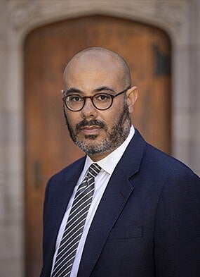 Man with brown skin, bald head, beard and glasses wearing a suit.