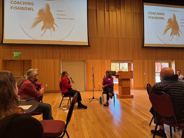 People sitting in chairs and having a discussion with mics and a screen behind them.