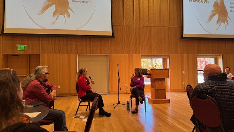 People sitting in chairs and having a discussion with mics and a screen behind them.