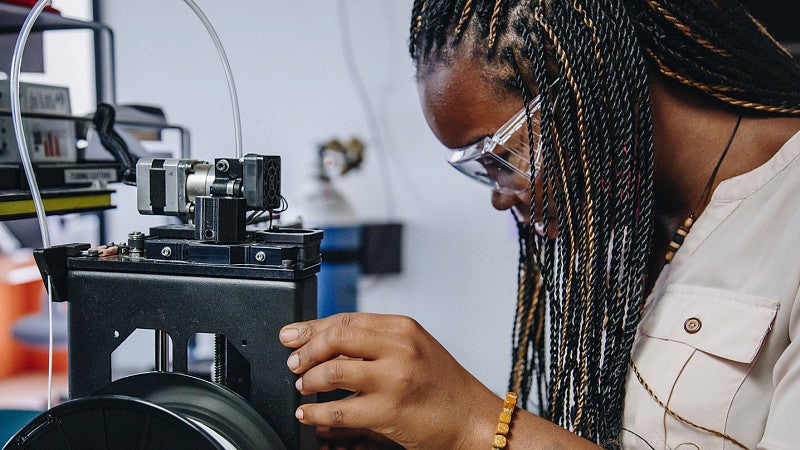 Student in lab coat with braids looking into equipment.